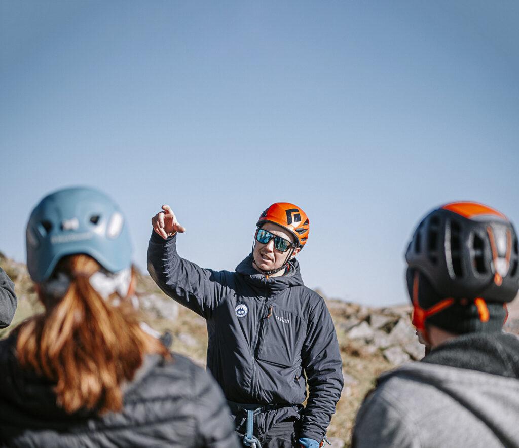 A man pointing whilst speaking to a group of people in rock climbing equipment