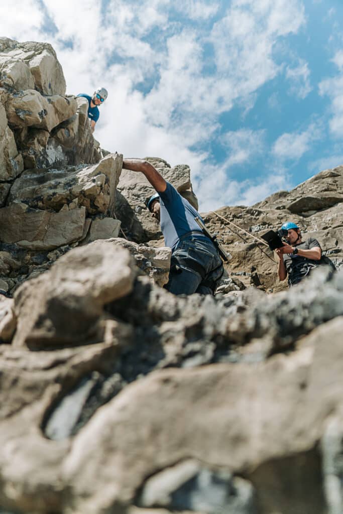 A rocky ledge and blue skies above. Amar Latif climbs the rock face whilst being filmed by a cameraman