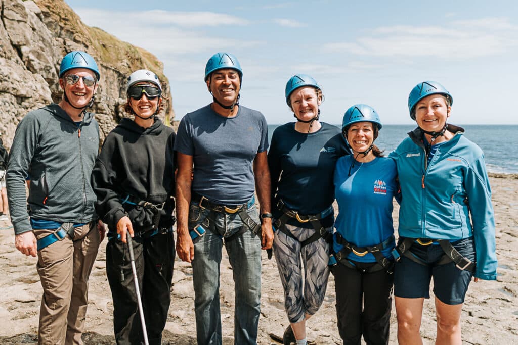 A group of six people stand on a grey rocky ledge smiling. They all wear blue climbing helmets. They include Nick Durrent from Acorn Adventure travel, Verity Church from Land & wave, Amir Latif OBE known as The Blind Adventurer, Sophie Reed from Activities Industry Mutual, Sam Baker from Dorset Blind Association and Rosie Tanner from Land & Wave