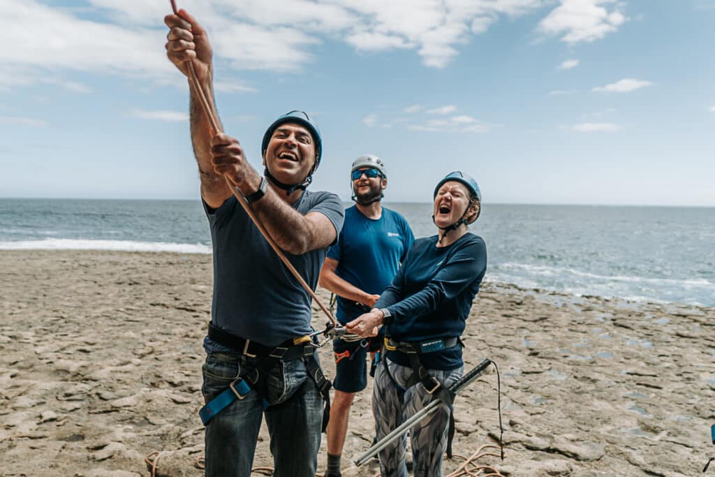 Amar Latif and his sighted guide, Sophia Reed belay a climber. They hold a rope. Alex Mortimer is an outdoor instructor who assists the pair from behind. They all stand on a rocky grey ledge with the sea in the background