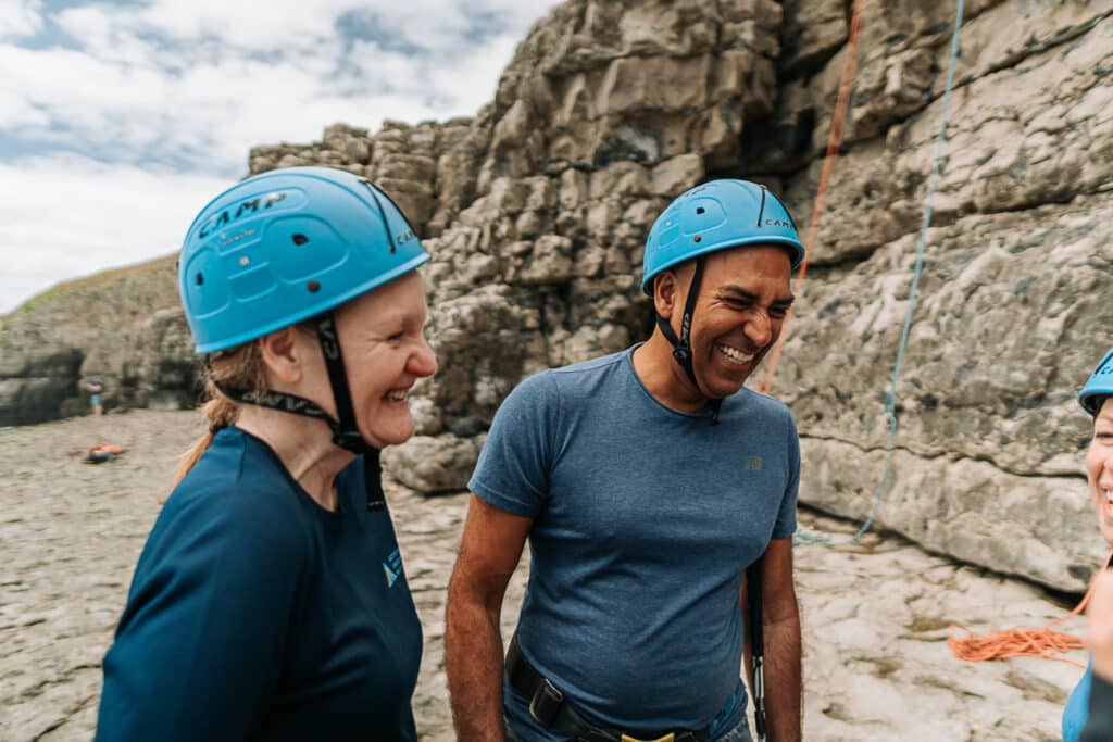 Amir Latif OBE laughs with Sophia Reed at the foot of a climbing route. They both wear blue climbing helmets and are smiling