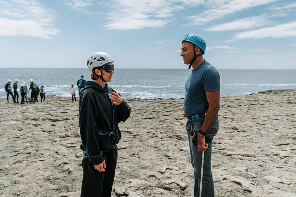 Amar Latif and outdoor instructor, Verity, stand chatting on a rocky grey ledge in the sunshine. The sea is in the background. Verity wears a white climbing helmet and Amar wears a blue climbing helmet