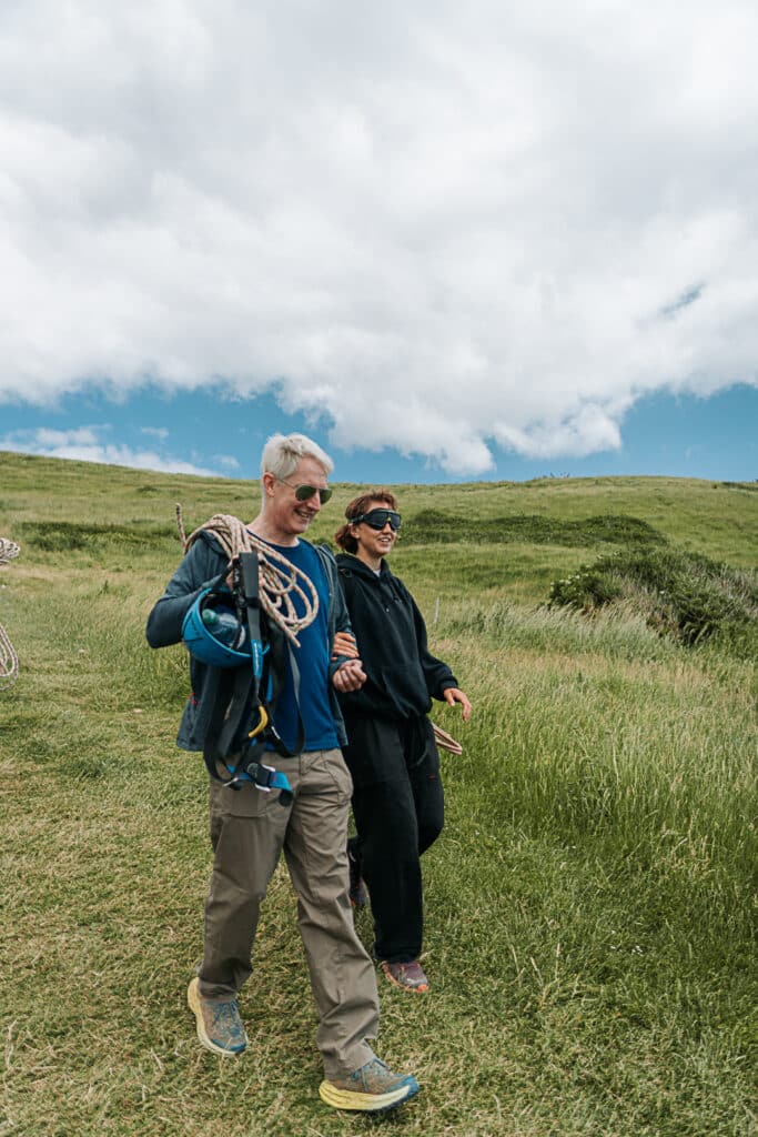 A grey haired man acts as a sighted guide for a person who is wearing blckout goggles. They are walking down a steep green hill in the countryside