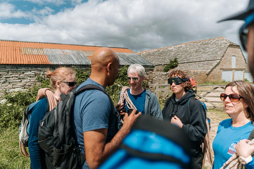 A group of four people stand outside some grey farm buildings chatting. They are holding climbing ropes and wearing backpacks. One person is wearing a pair of blackout goggles.