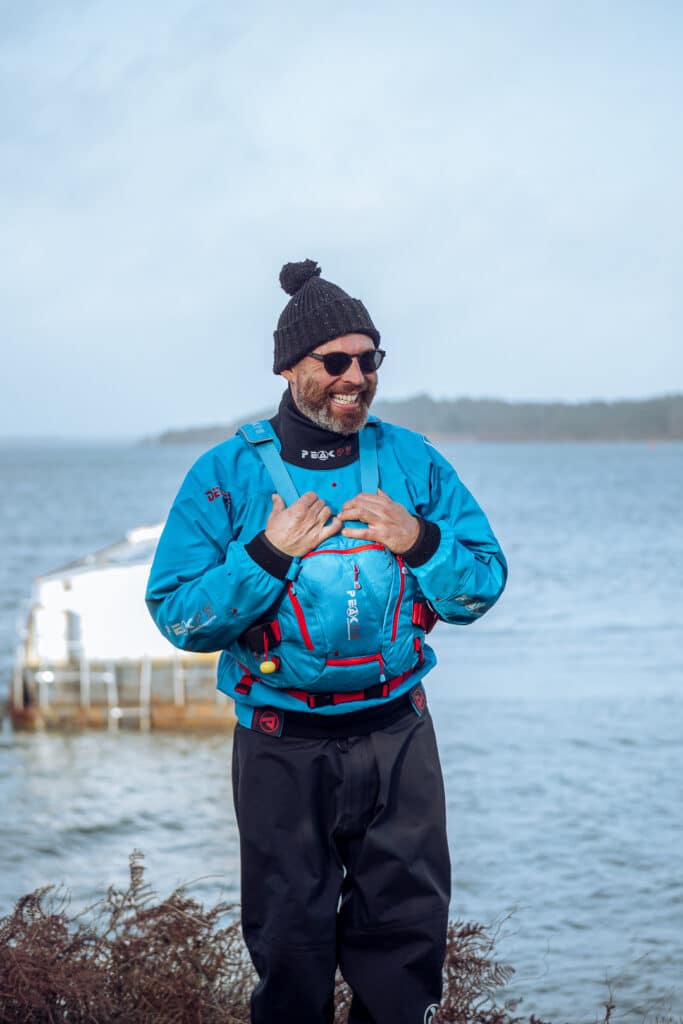 A man stands next to the sea wearing a drysuit, buoyancy and a woolly hat.