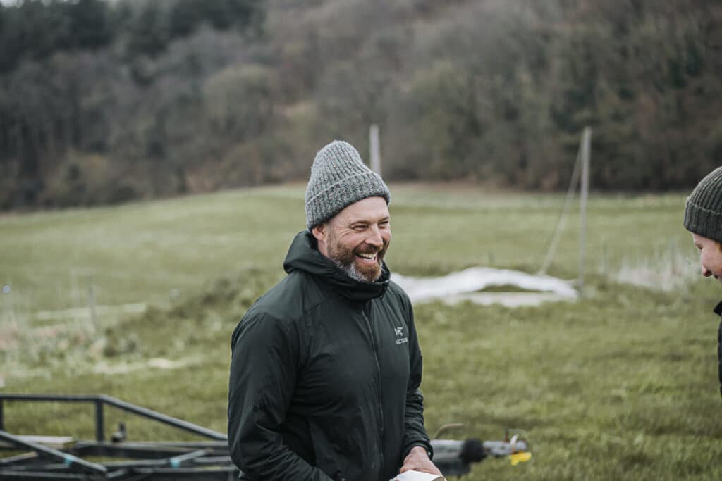 A man with AdHD laughs as he stands in a green countryside field wearing a woolly hat and black jacket